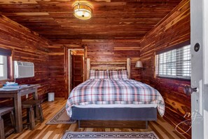 A wooden cabin bedroom with a bed, a desk, and a window.