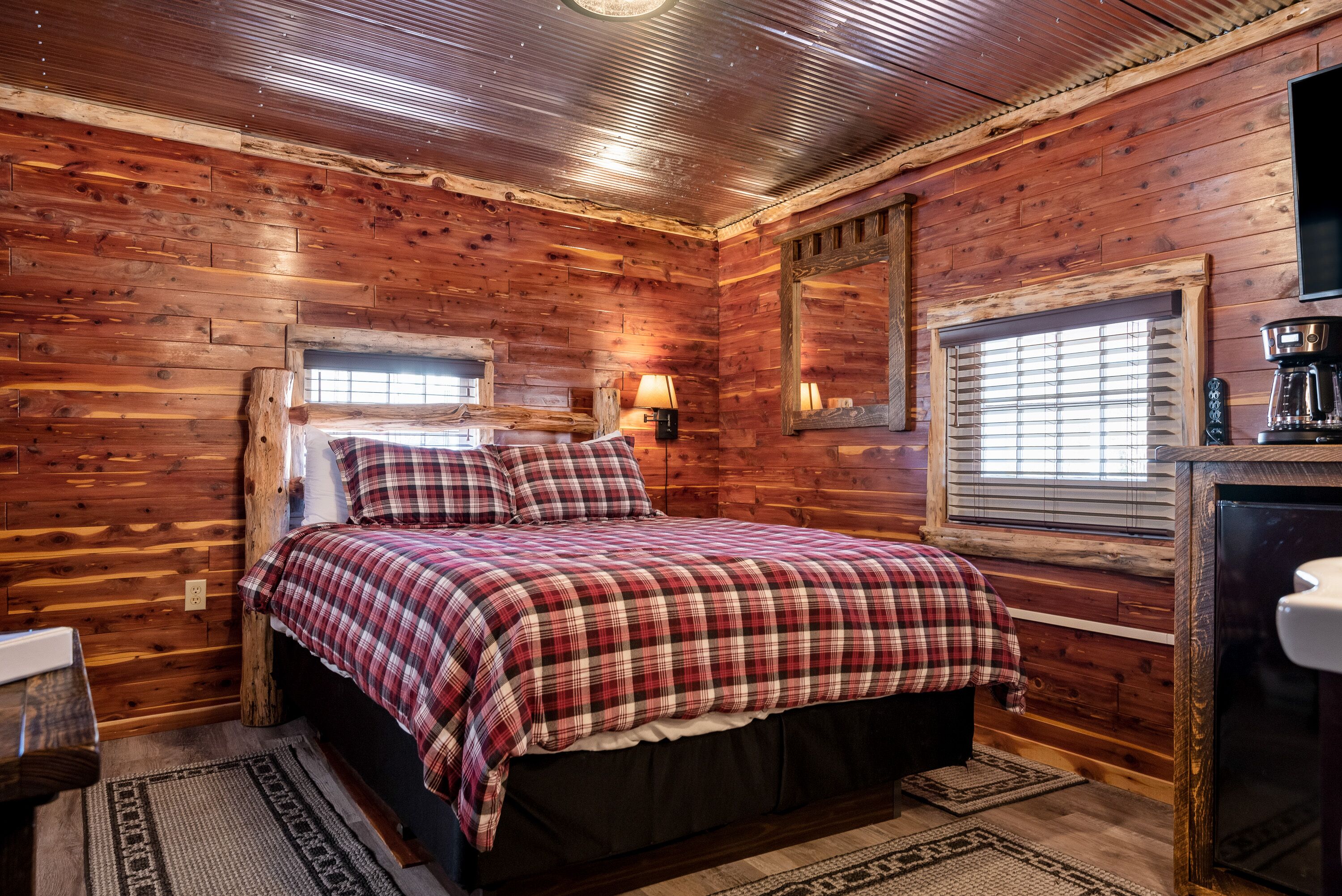 A cozy log cabin bedroom with a large bed, wooden walls, a window with blinds, and a coffee maker on a wooden table.