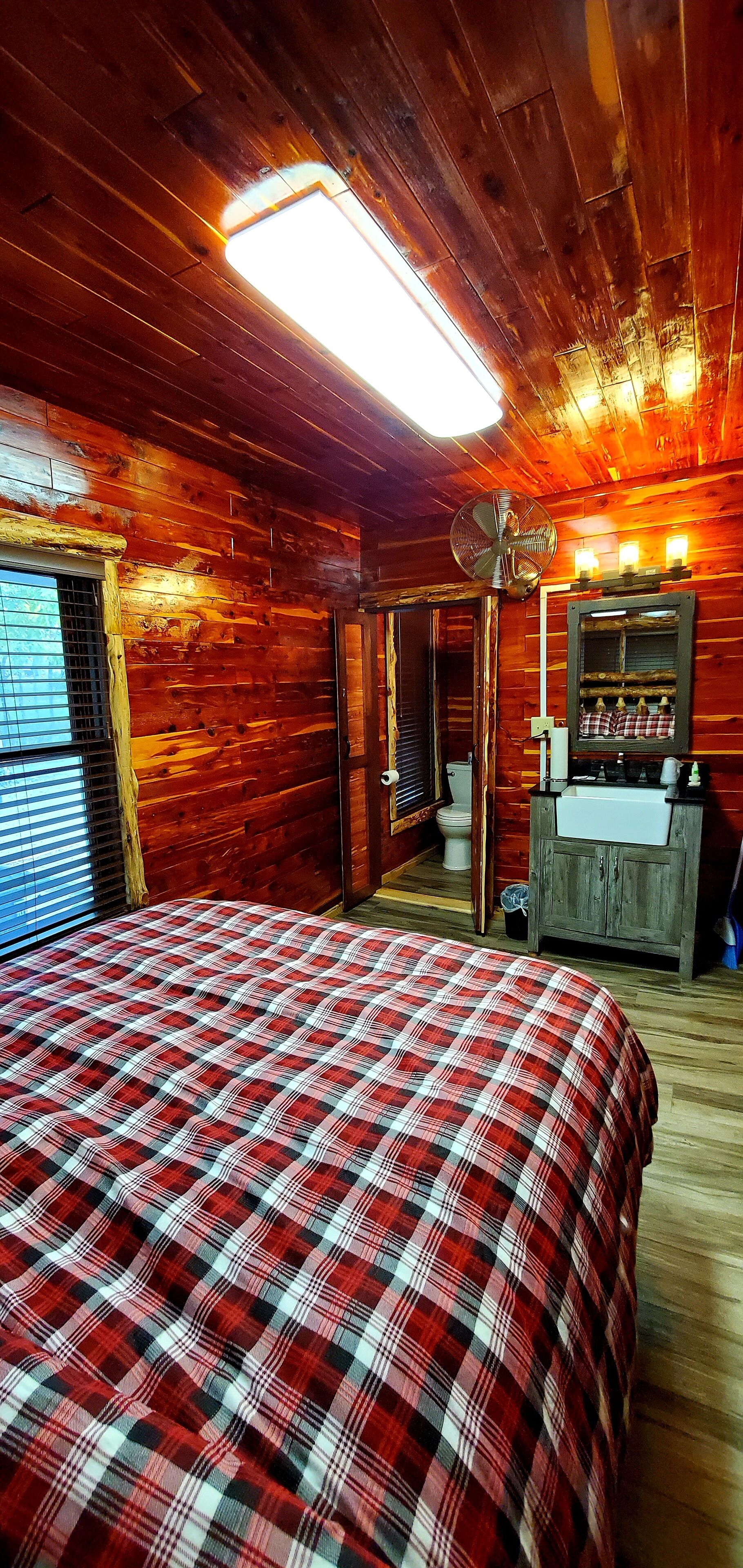 A wooden cabin bedroom with a plaid bedspread, a fan, a mirror, and a window with blinds.