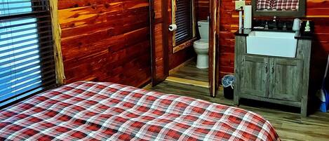 A wooden cabin bedroom with a plaid bedspread, a fan, a mirror, and a window with blinds.