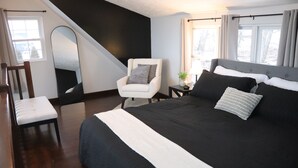 A bedroom with a large bed, a white armchair, a small table with a lamp, and a window with curtains.