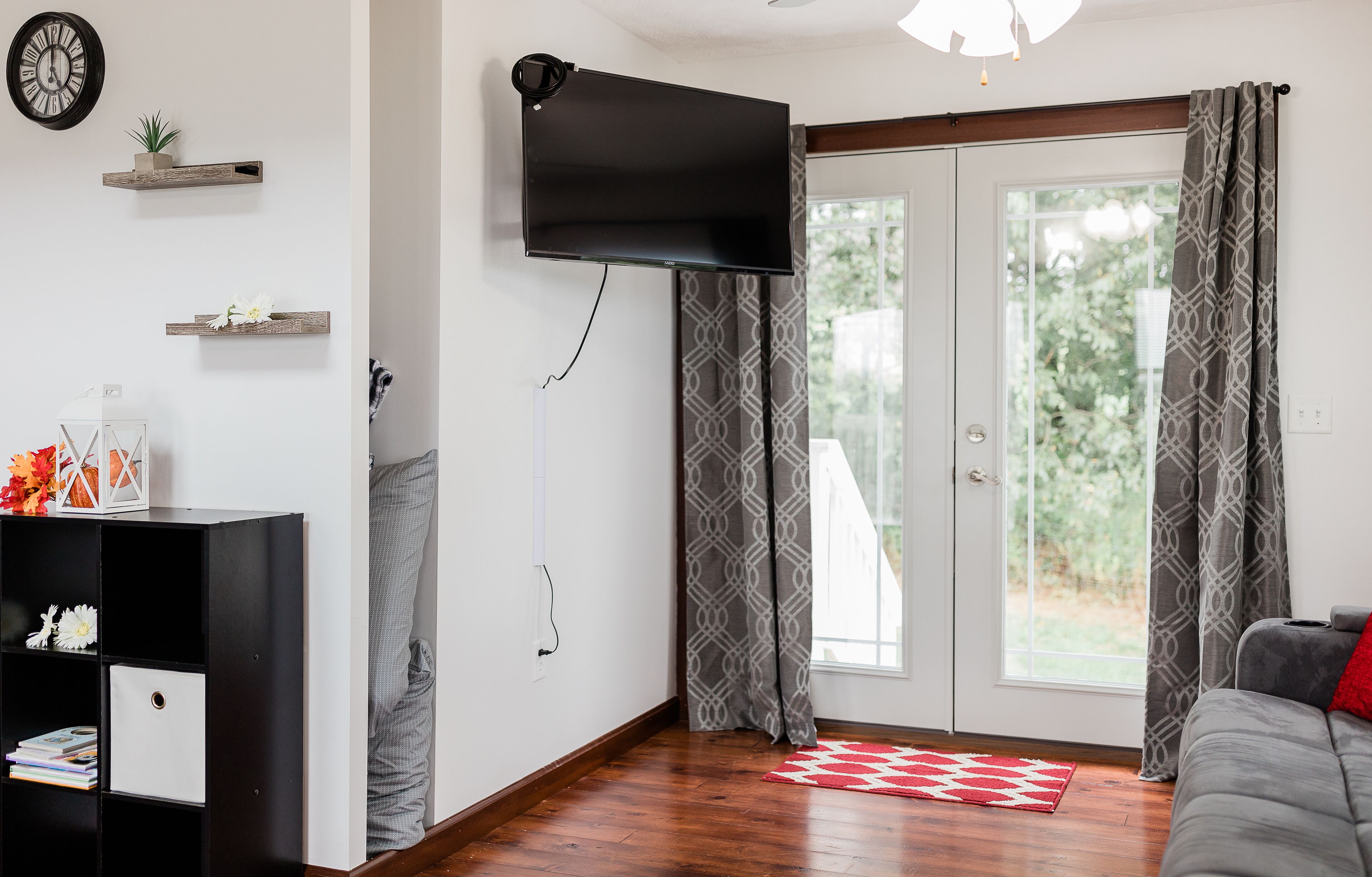 A living room with a flat-screen TV mounted on the wall, a wooden floor, a sofa, a red and white rug, and a sliding glass door with patterned curtains.