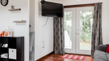 A living room with a flat-screen TV mounted on the wall, a wooden floor, a sofa, a red and white rug, and a sliding glass door with patterned curtains.
