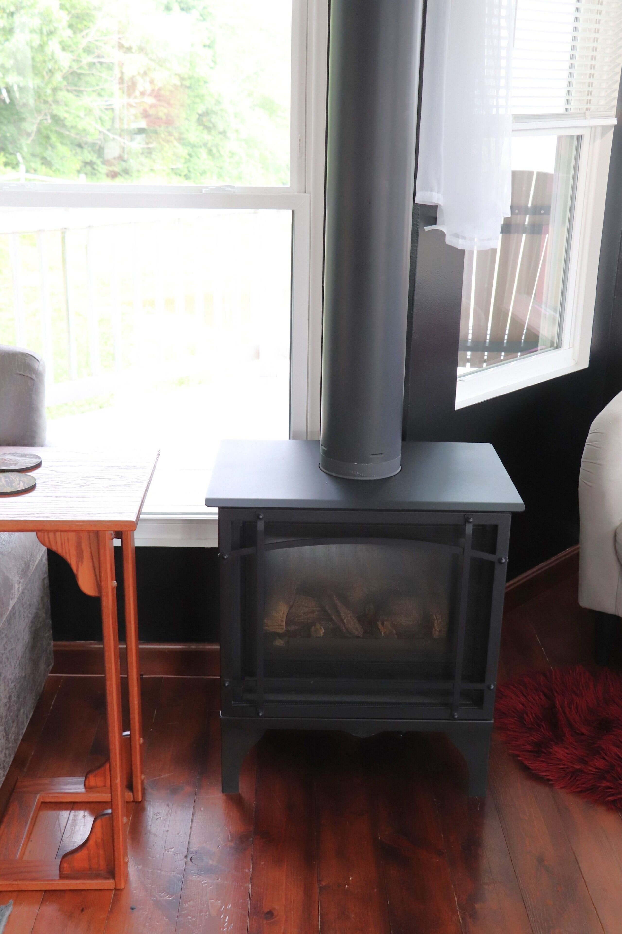 A wood-burning stove with a glass door, a wooden table with a chair, and a window with blinds.