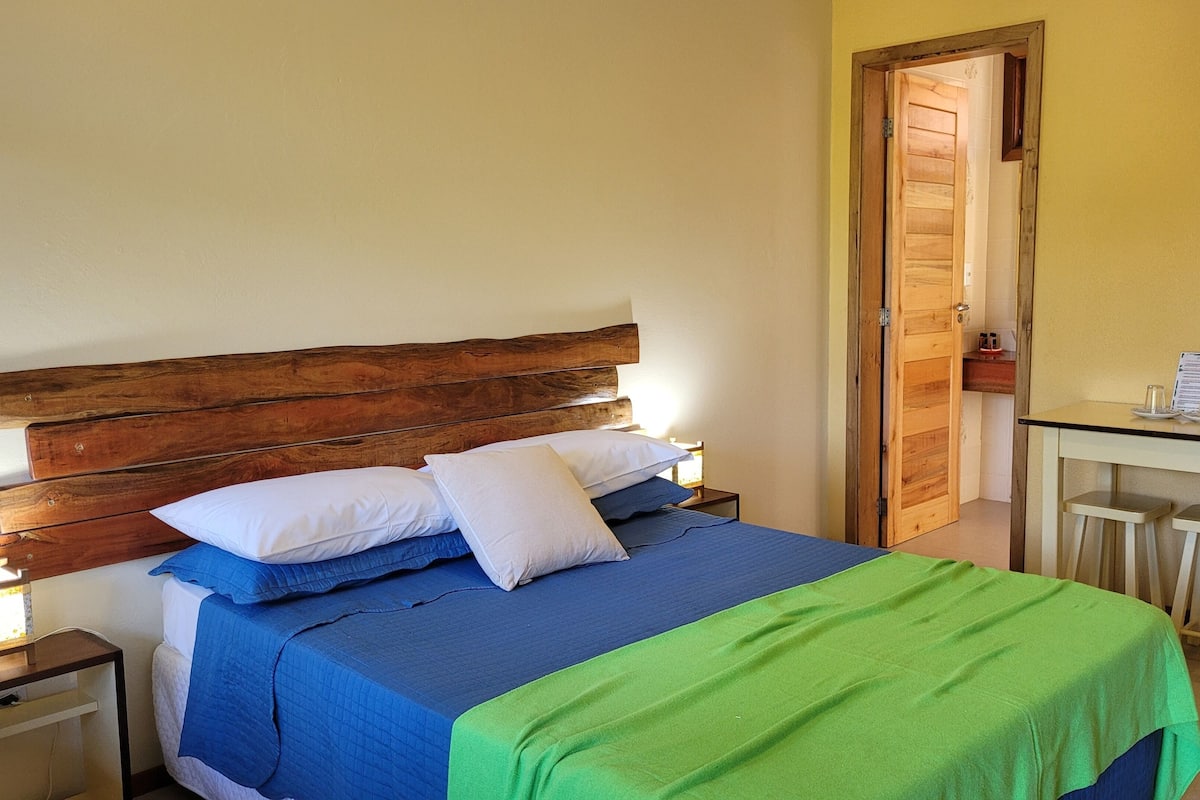 A bed with a wooden headboard, blue and green bedding, and white pillows. There is a small table with two chairs and a menu on it.