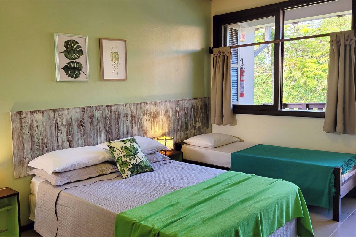 A room with two beds, a wooden headboard, framed botanical prints, a window with curtains, and a nightstand with a lamp.