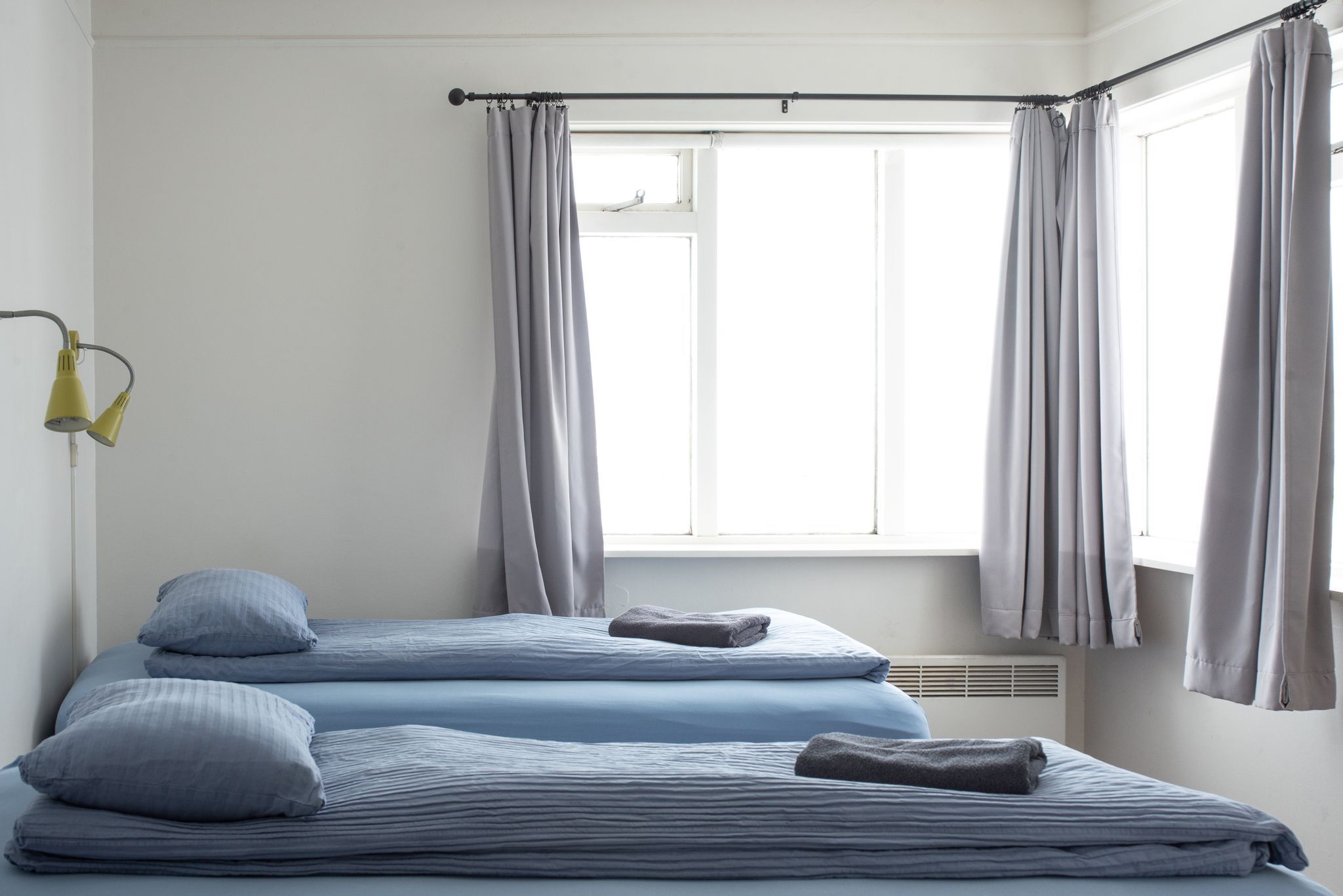 Une chambre avec deux lits, une fenêtre avec des rideaux, un radiateur et une lampe fixée au mur.