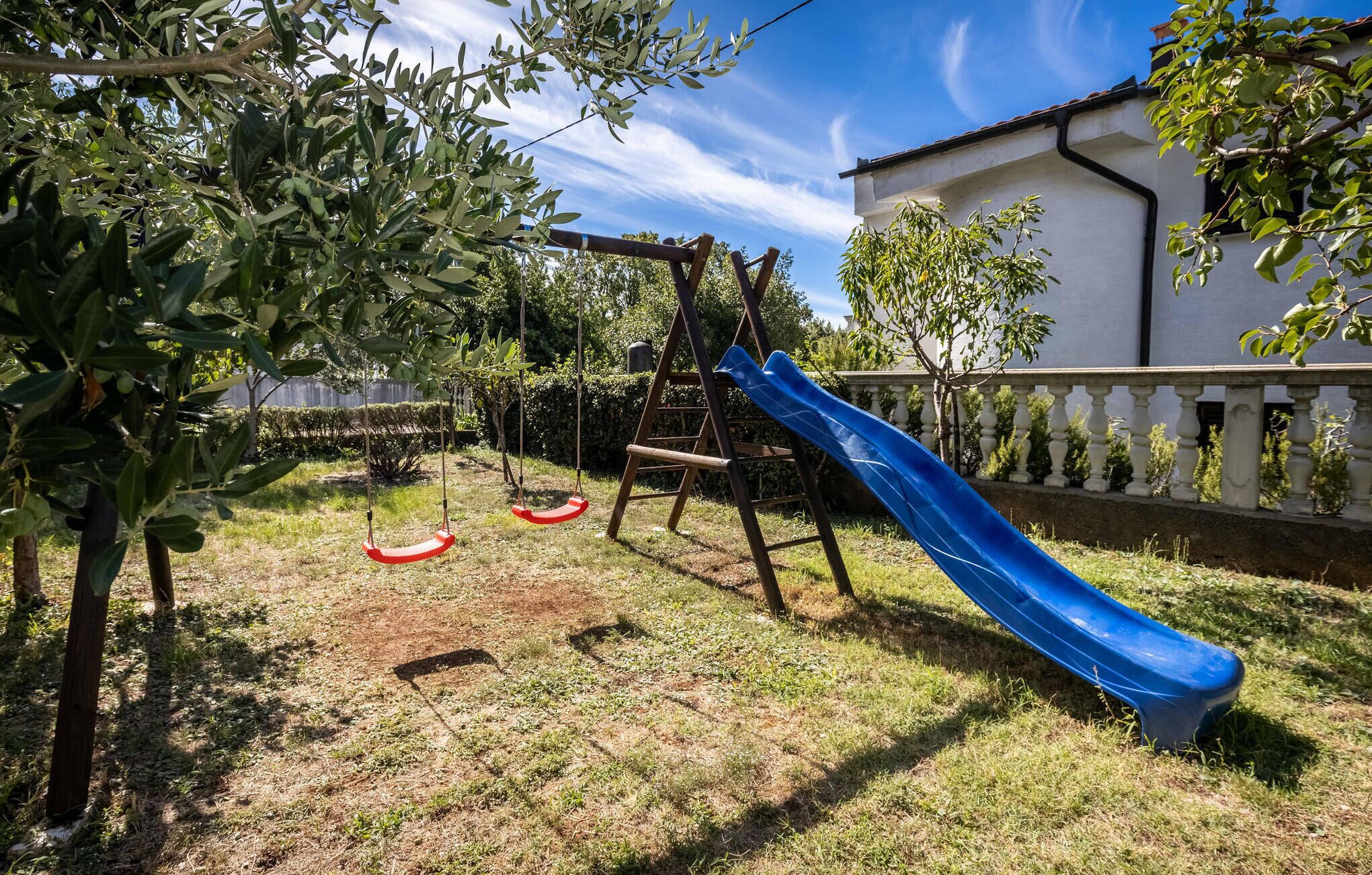 A playground with a slide and swings, surrounded by trees and a white building.