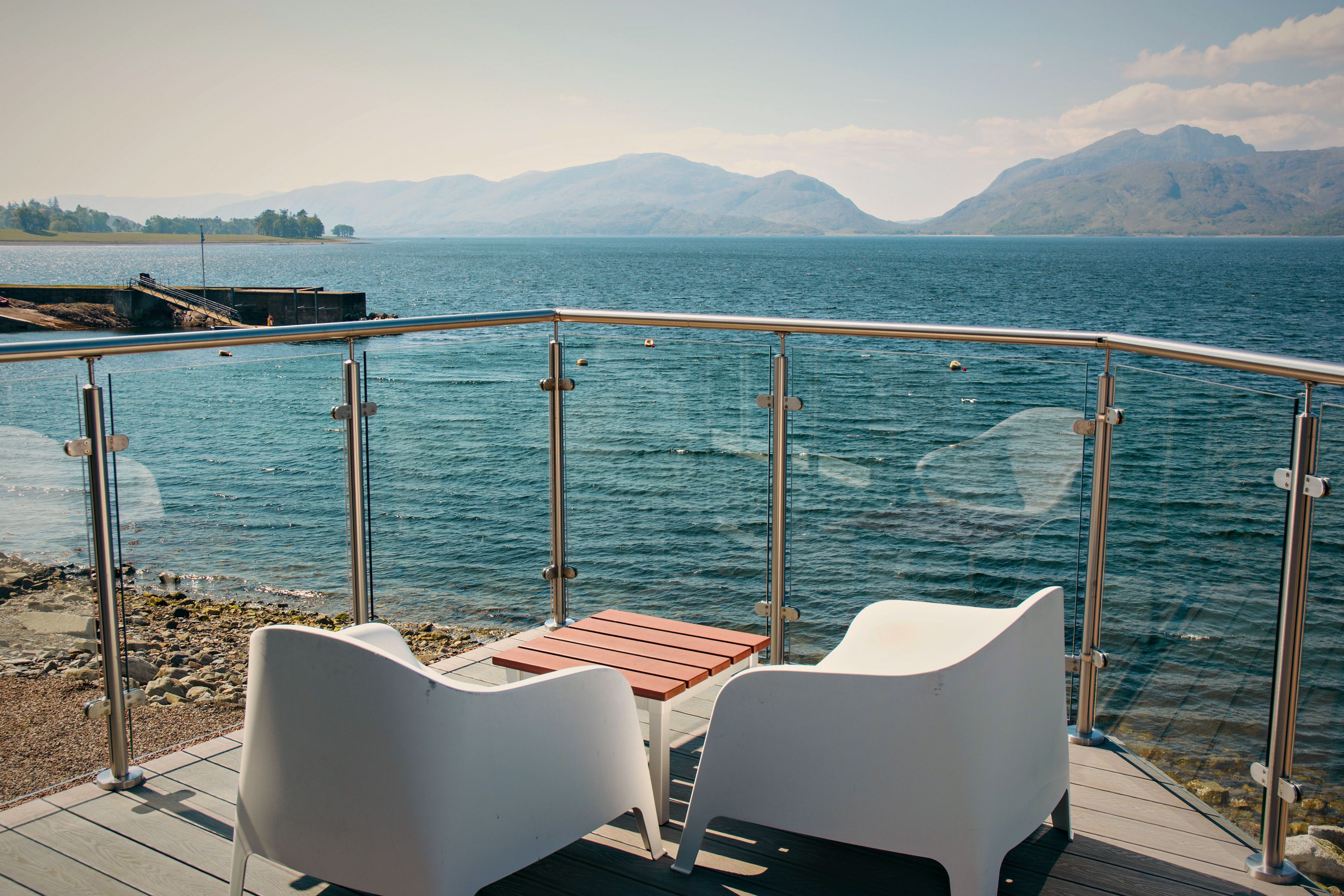 Une terrasse avec deux chaises blanches et une petite table, d’où l’on a vue sur une étendue d’eau, avec des montagnes à l’horizon.