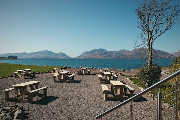 Een picknickplaats aan het meer met houten tafels en banken, een grindpad en een uitzicht op bergen in de verte.