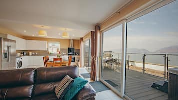 A modern living room with a leather sofa, a dining area, and a kitchen. There is a large sliding glass door leading to a balcony with a view of the water.