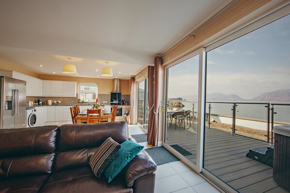 A modern living room with a leather sofa, a dining area, and a kitchen. There is a large sliding glass door leading to a balcony with a view of the water.