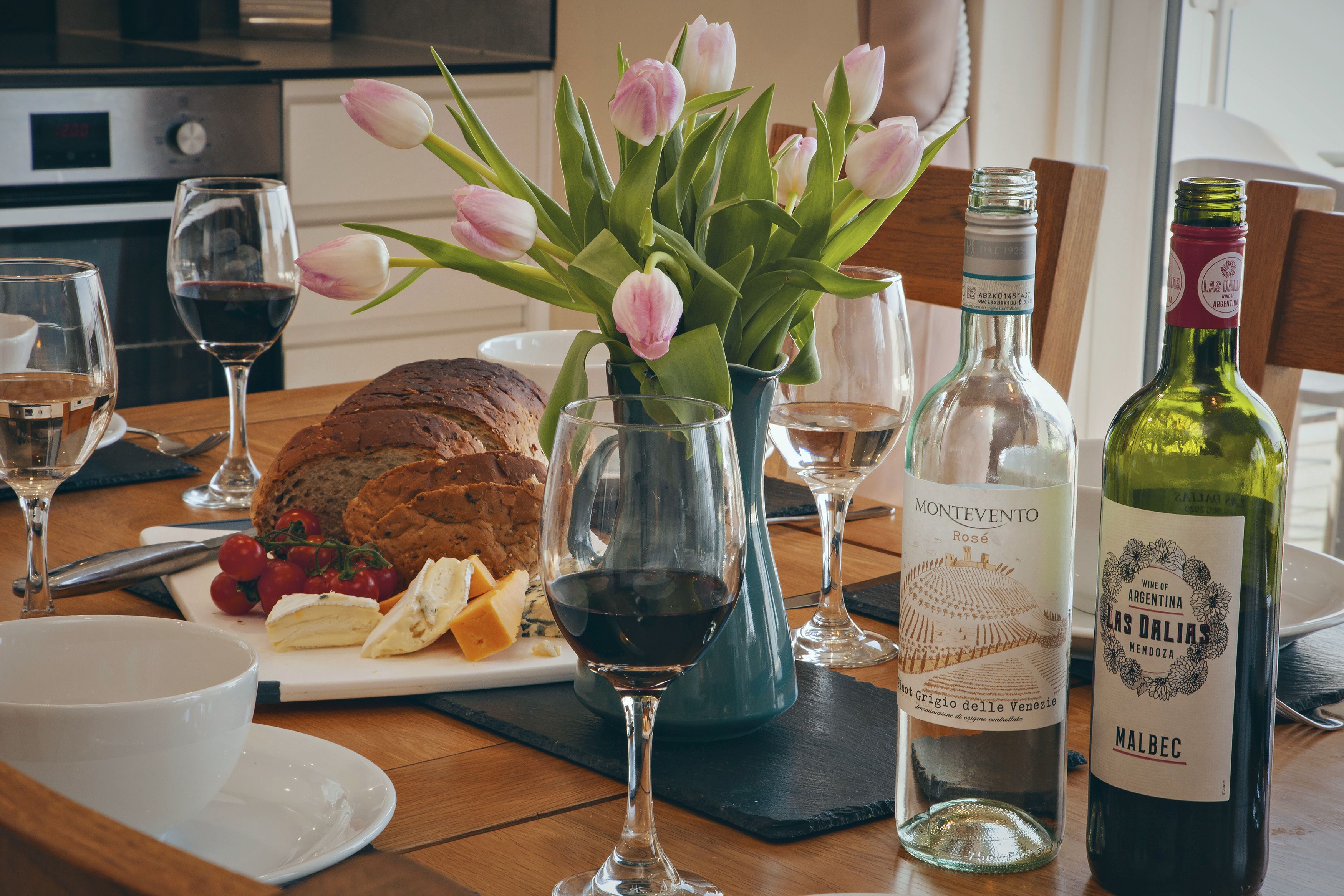 A dining table with a loaf of bread, cheese, tomatoes, and a glass of red wine. There are also tulips in a vase, a bottle of wine, and a glass of white wine.