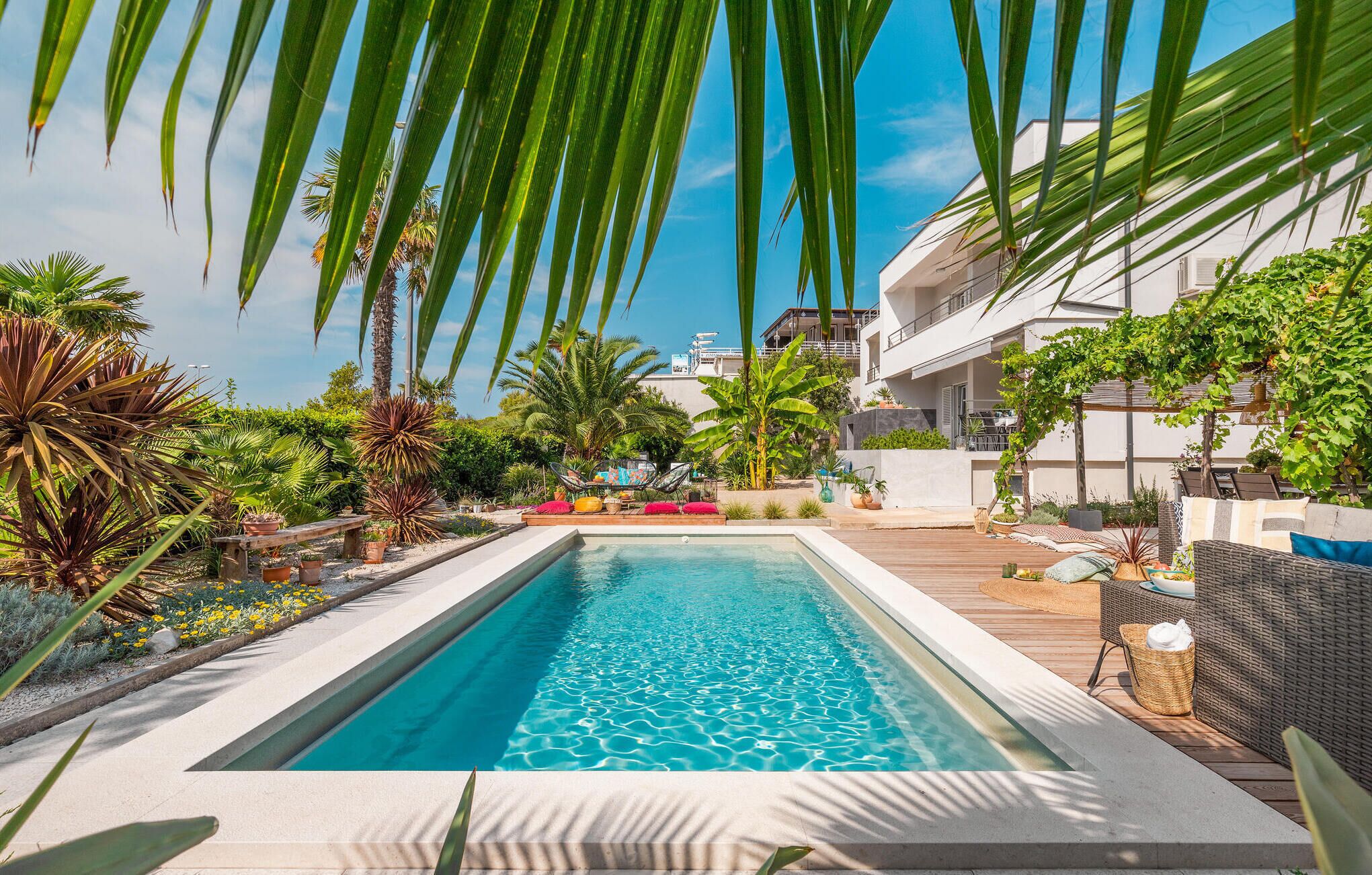 A rectangular swimming pool with clear blue water, surrounded by a wooden deck and various tropical plants.