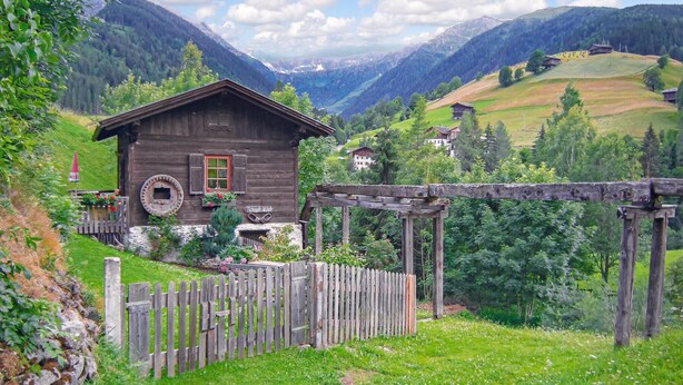 Ein rustikales Holzhäuschen mit Holzzaun, umgeben von üppiger Vegetation und Bergen im Hintergrund.