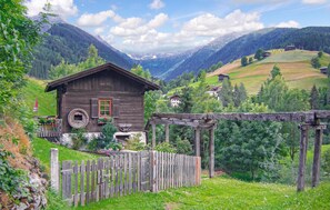 Ein rustikales Holzhäuschen mit Holzzaun, umgeben von üppiger Vegetation und Bergen im Hintergrund.