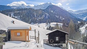 Eine Holzhütte mit schneebedecktem Dach und einer Holzzahlung, eingebettet in eine verschneite Berglandschaft unter klarem Himmel.