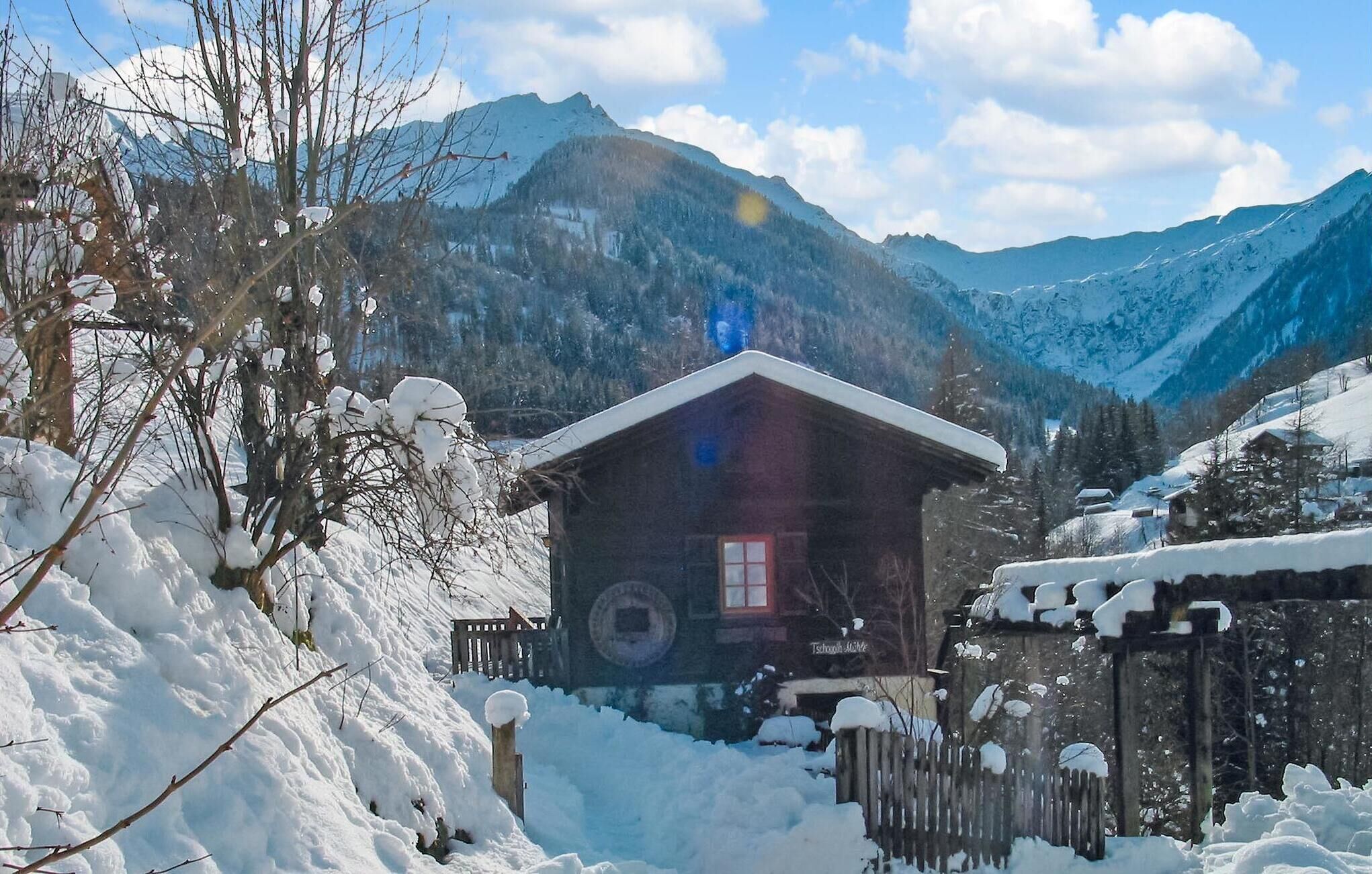 A wooden cabin with a red door and windows, surrounded by snow-covered trees and mountains.