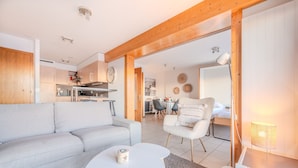Una sala de estar moderna con un sofá, una mesa de centro y un sillón. Se aprecia una cocina con armarios y electrodomésticos al fondo.