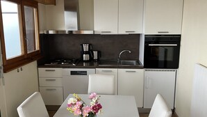 Eine moderne Küche mit weißen Schränken, einer Edelstahl-Dunstabzugshaube, einer Kaffeemaschine und einem Backofen. Ein Esstisch mit Stühlen und eine Blumenvase sind ebenfalls vorhanden.