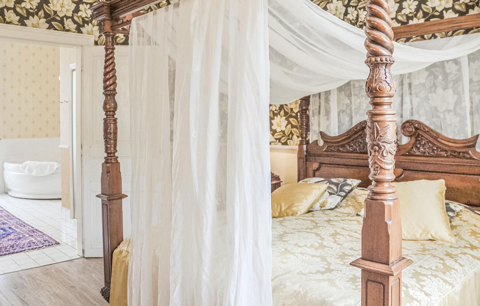 A bedroom with a four-poster bed, a white canopy, floral wallpaper, and a white toilet in the adjacent room.