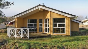 Une maison en bois d’un seul étage, dotée d’un toit à pignon, de grandes fenêtres et d’une terrasse avec une balustrade blanche.