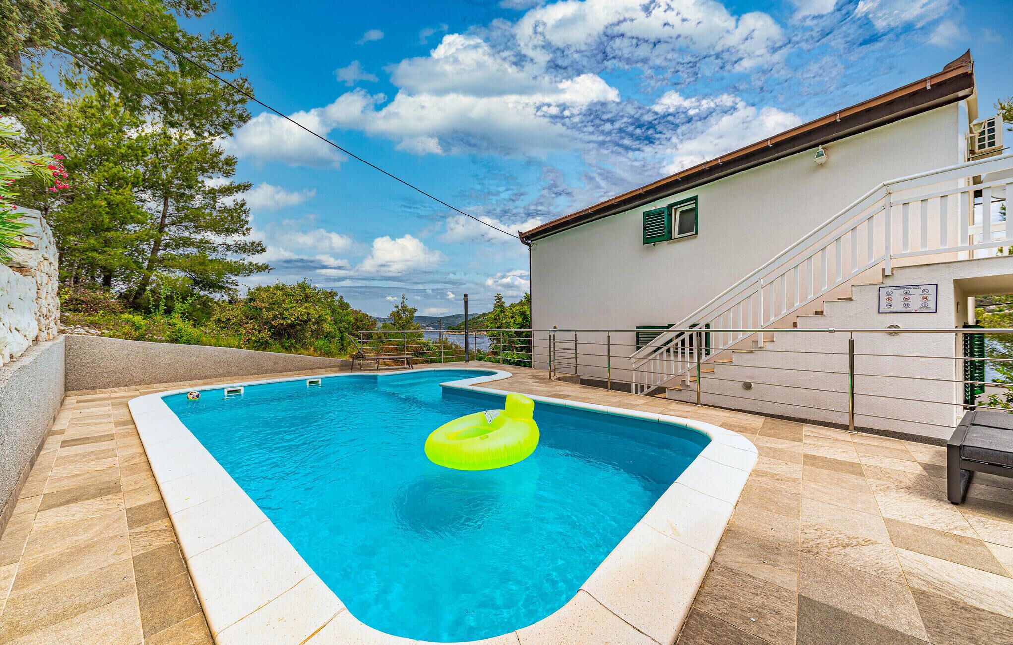 Ein Schwimmbecken mit einem grünen Luftring, einer terrasse mit Steinplatten und ein weißes Gebäude mit einer Treppe und einem Schild.