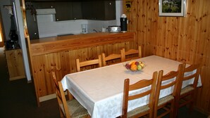Un coin repas en bois avec une table dressée pour quatre personnes, un panier de fruits et une cuisine en arrière-plan.