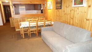 Une pièce chaleureuse avec une table et des chaises en bois, un canapé gris et un coin cuisine à l’arrière-plan.