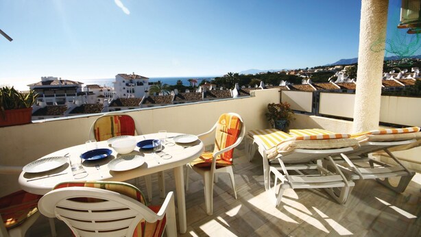 A terrace with a table set for dining, white chairs, and a view of the sea and buildings. - Beautiful apartment in Calahonda (Calahonda)