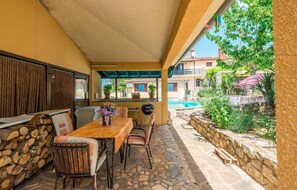 A covered outdoor dining area with a table and chairs, a grill, and a view of a pool and buildings in the background. - Awesome apartment in Porec (Porec)