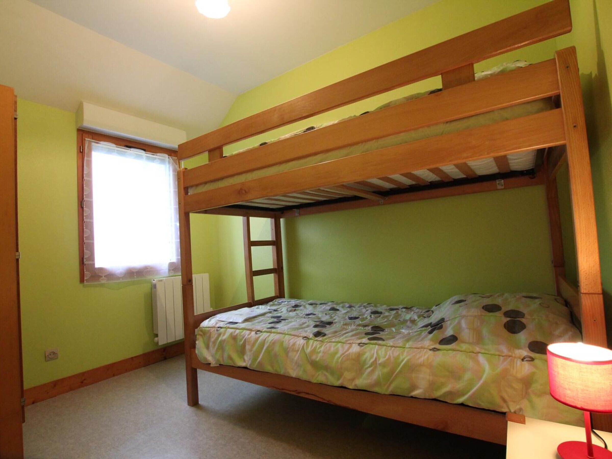 Un lit superposé dans une chambre aux murs verts, avec une fenêtre munie de stores et une lampe rouge posée sur une table de chevet.