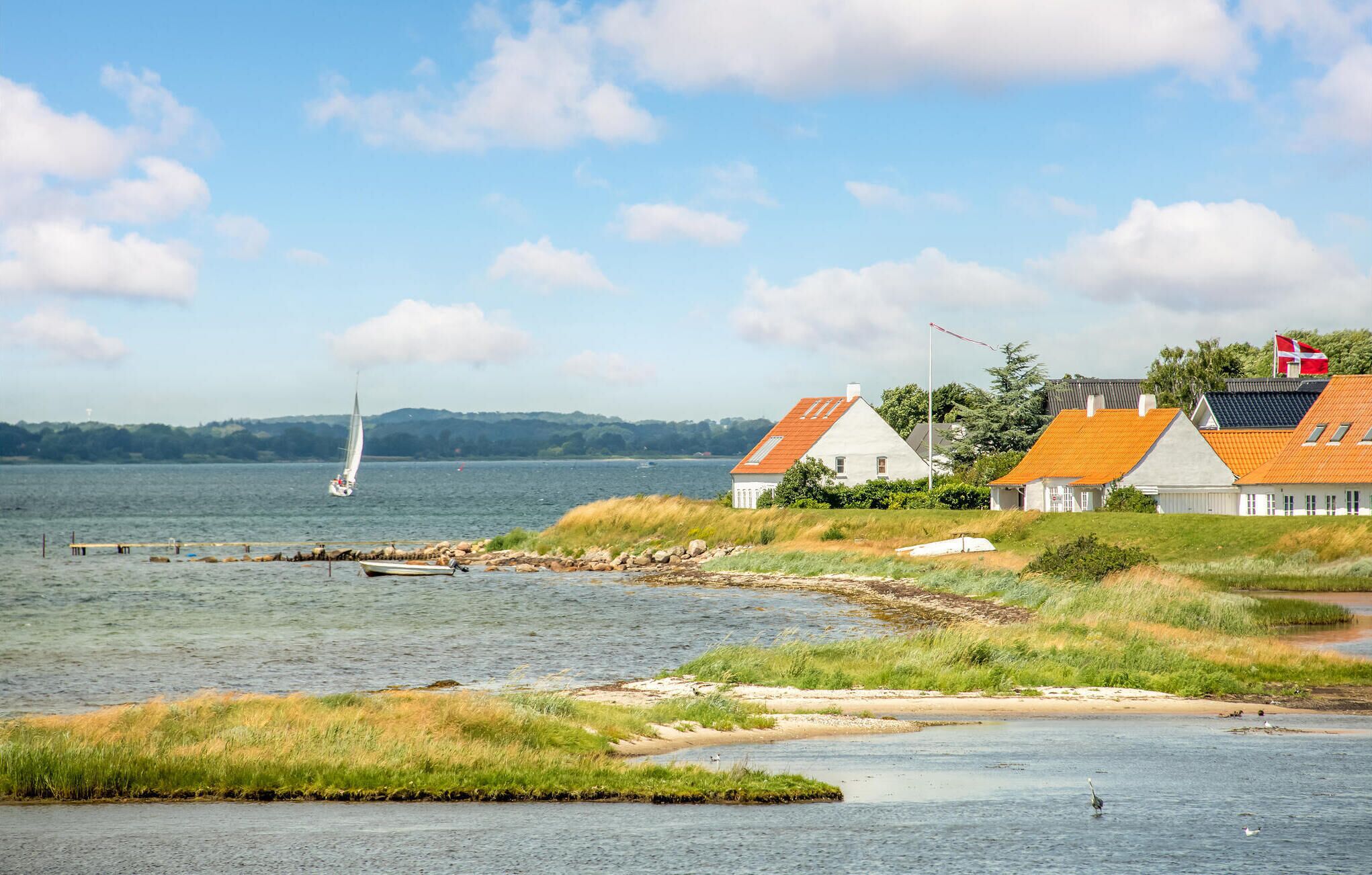 A coastal scene with a sailboat on the water, a rowboat near the shore, and houses with orange roofs.