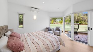 A modern bedroom with a large bed, two chairs, and a view of a garden through glass doors. - THE LODGE - TENNIS,LGE SPA,WOOD FIRE, (Anglesea)