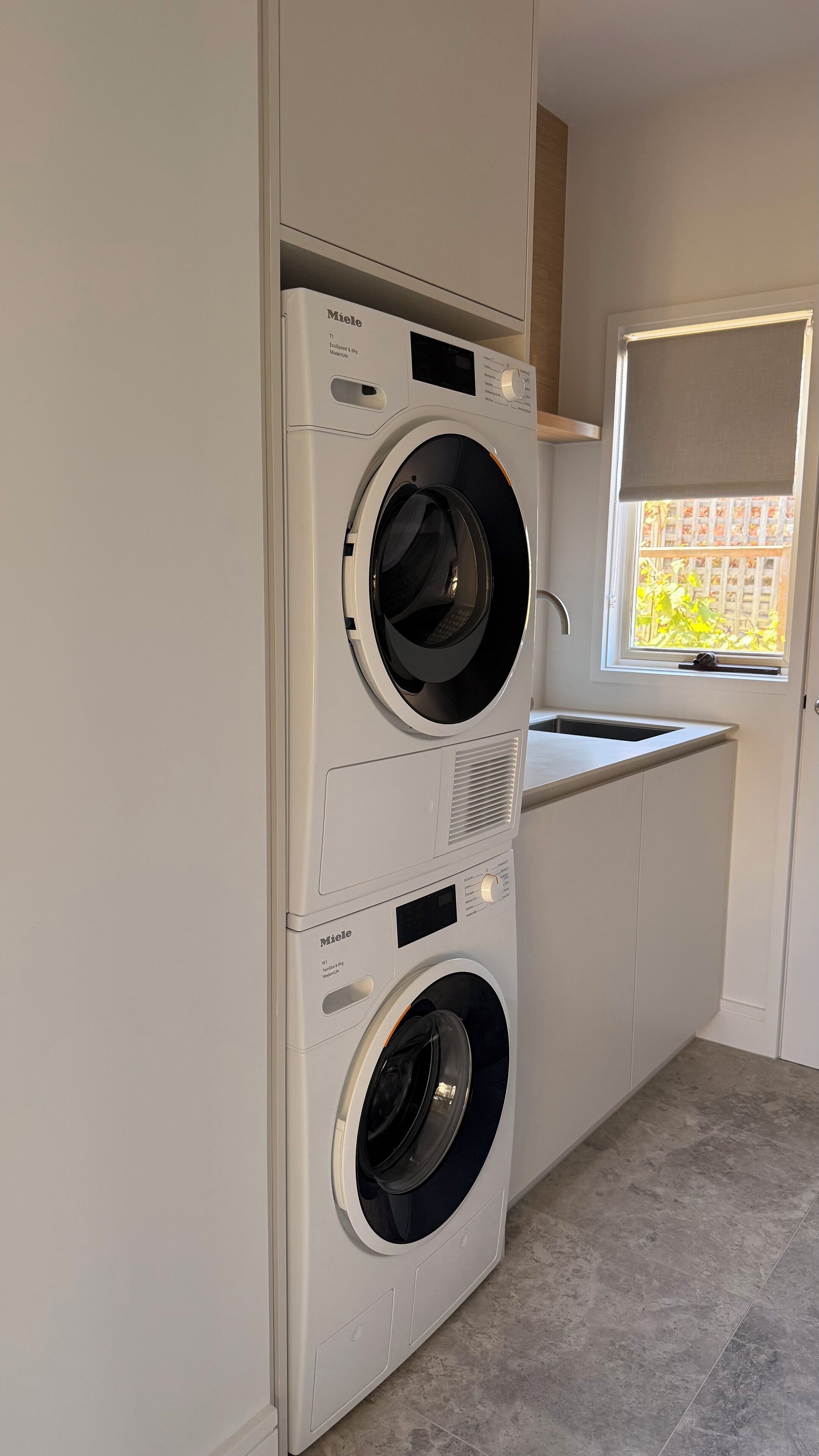 A compact, modern laundry room with two white front-loading washing machines, a small window with a view of greenery, and a built-in sink.