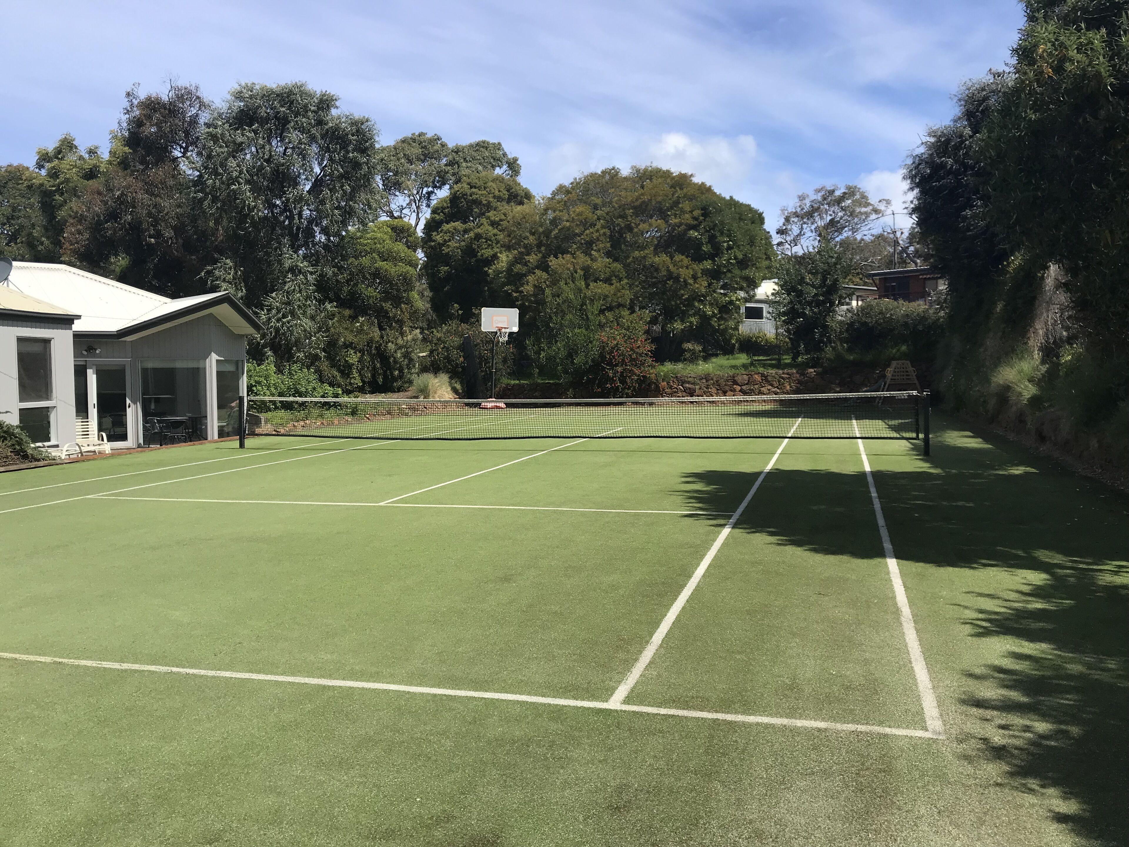 Een tennisbaan met een net, omgeven door bomen en een gebouw met een overdekte zithoek.