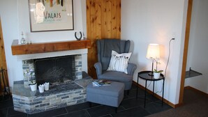 Ein Wohnzimmer mit einem Kamin aus Stein, einem grauen Sessel, einem kleinen Tisch mit einer Lampe und einem gerahmten Kunstwerk darüber.