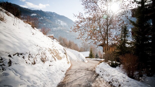 Ein verschneiter Weg führt zu einem Gebäude mit geneigtem Dach, umgeben von Bäumen und Bergen im Hintergrund.