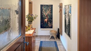 Un passadís amb un armari de fusta, un ram de flors en un vas, un tapís amb escenes de cérvols, un petit tamboret i un quadre encimbellat a la paret.