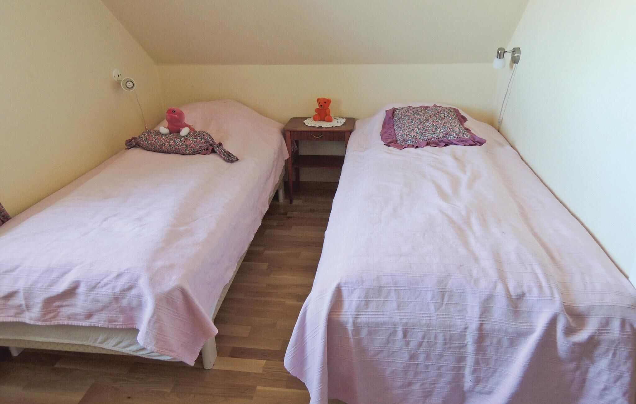 A small, cozy room with two single beds, a nightstand with a teddy bear, and a wall-mounted lamp.