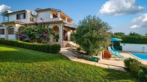 Ein zweistöckiges Haus mit Swimmingpool, einem Garten mit verschiedenen Pflanzen und einem blauen Sonnenschirm.