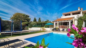 Ein Schwimmbecken mit klarem, blauem Wasser, umgeben von einer Terrassenanlage mit Liegestühlen und einem Trampolin.