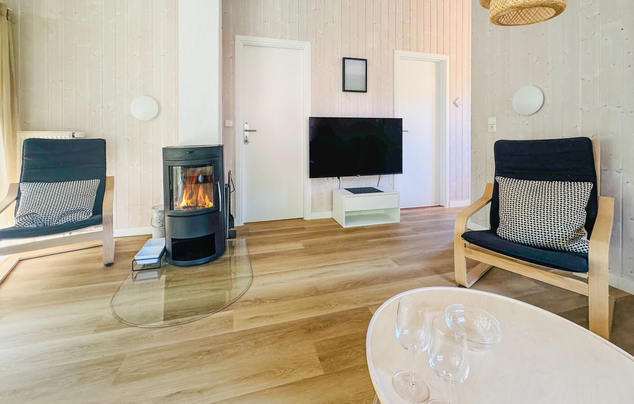 A cozy living room with a wood-burning stove, two black chairs with patterned cushions, a flat-screen TV on a white stand, and a glass coffee table.