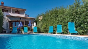 A swimming pool with blue chairs and umbrellas, a stone building with a tiled roof, and a dense green hedge in the background. - Stunning home in Pula (Pula)