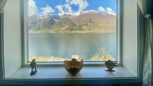 A window with a view of a lake and mountains, with two small figurines on the windowsill.