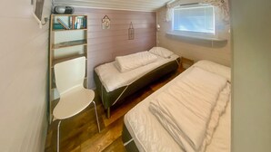 A compact room with two beds, a small white chair, and a shelf with books and a clock.