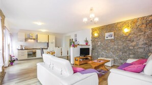 Una sala de estar moderna con sofás blancos, una mesa de centro y un mueble para la televisión. Se ve una cocina al fondo, con armarios blancos y una pared de piedra.
