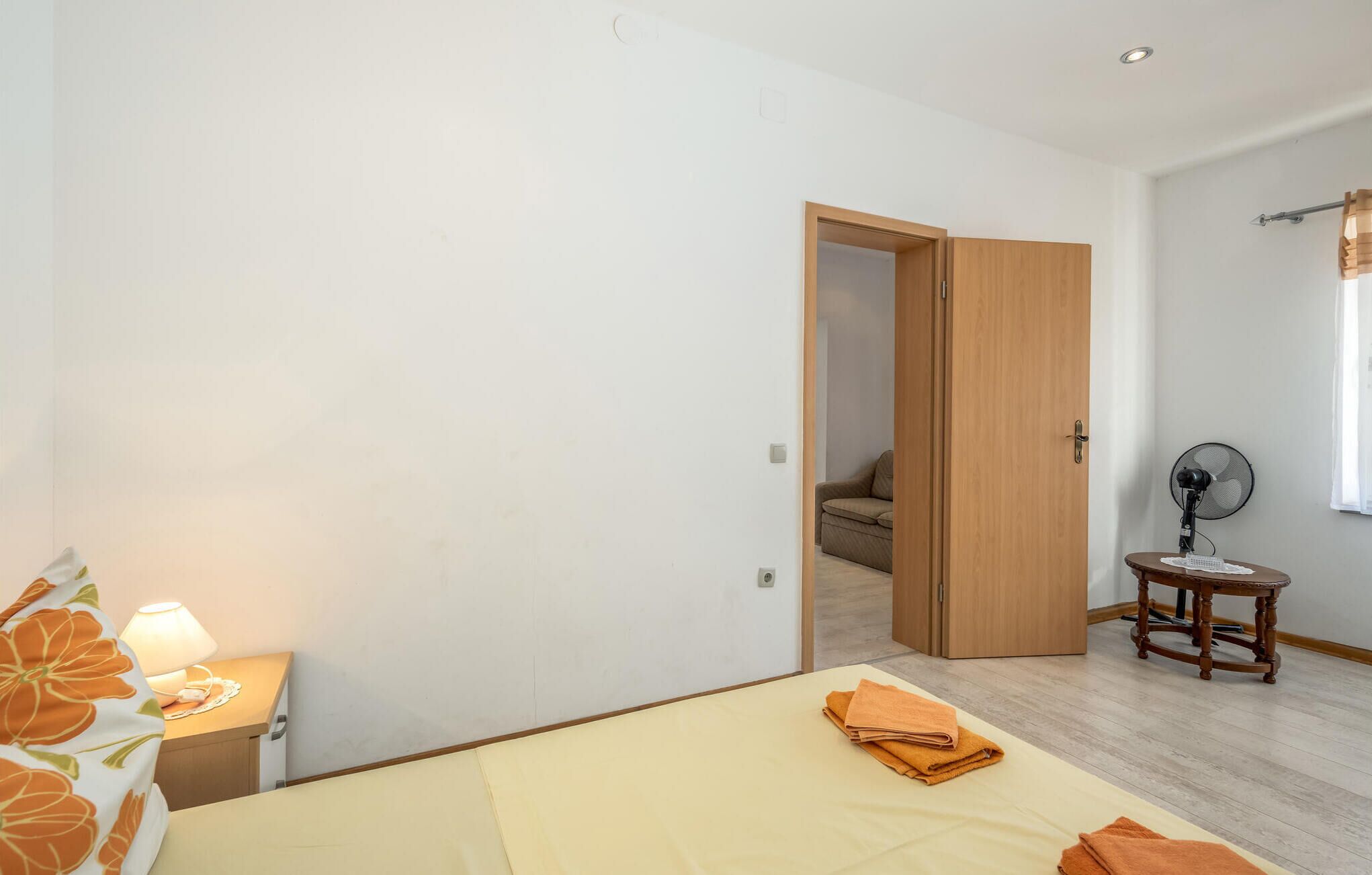 A bedroom with a bed, a nightstand, a fan, and a door leading to another room.