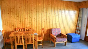 Een kamer met houten lambrisering, een eettafel met stoelen, een gestreept fauteuil en een blauwe poef.
