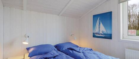 A small, single-bed room with white walls and a sloped ceiling. There is a painting of a sailboat on the wall, a window with a view of trees, and a white nightstand with a lamp.